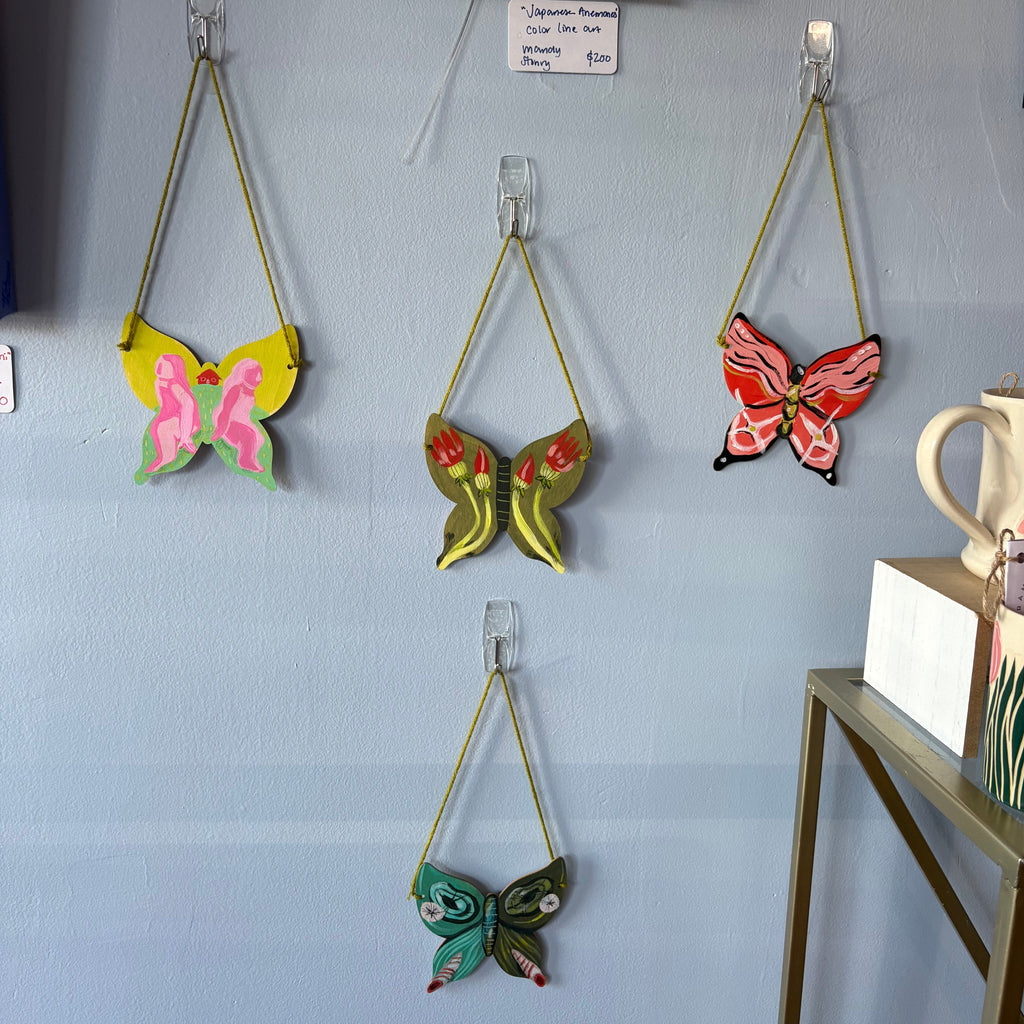Three colorful painted butterfly wall hangings suspended from clear clips on a light blue wall, with a small framed print and shelf visible in the room.