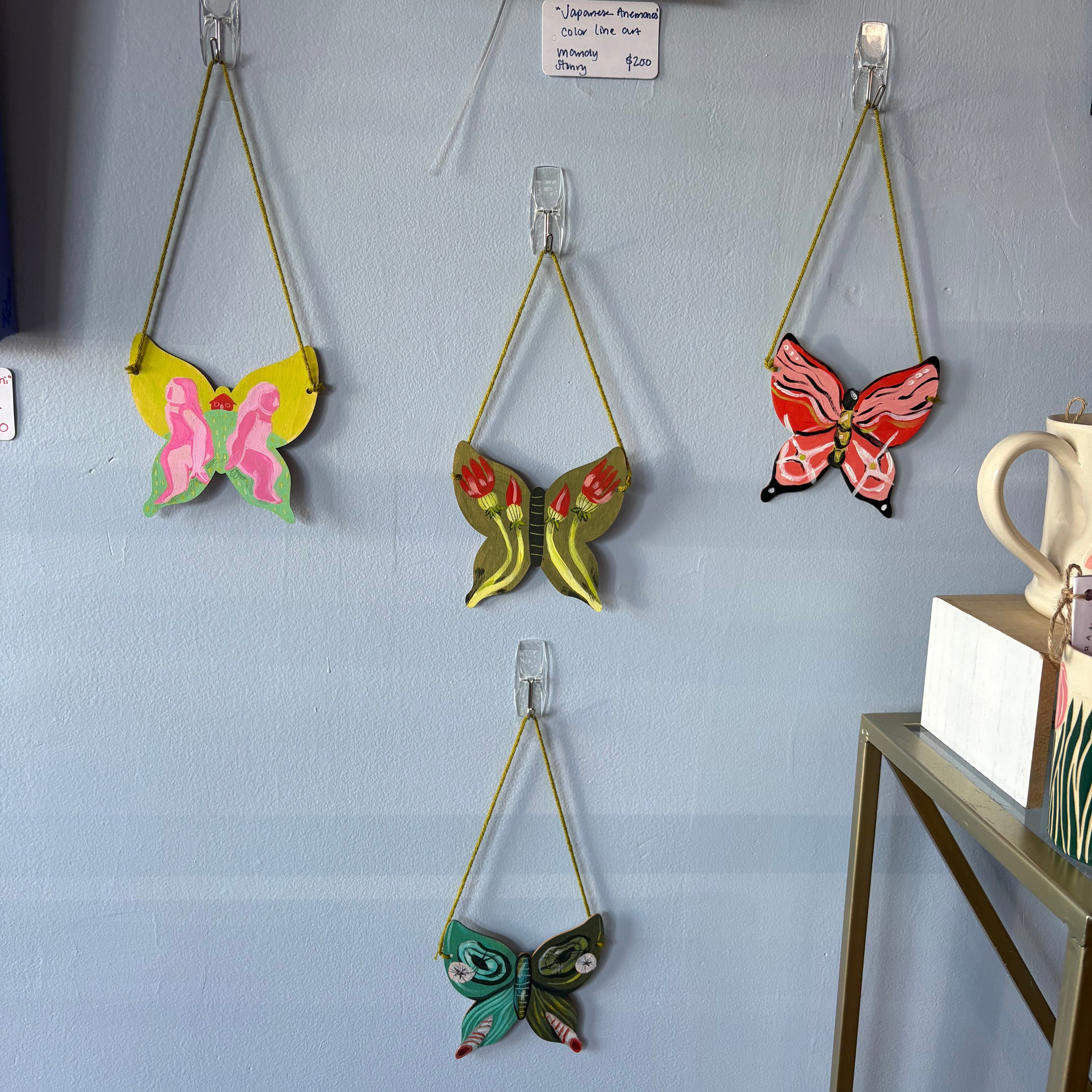 Three colorful painted butterfly wall hangings suspended from clear clips on a light blue wall, with a small framed print and shelf visible in the room.